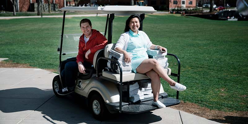 Two students riding a golf cart delivering the student newspaper around campus