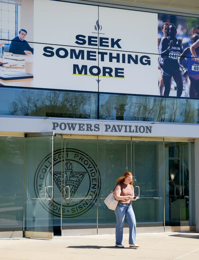 PC student walking out of Powers Pavilion doors of Slavin with a Seek Something More image showing out of the window from inside